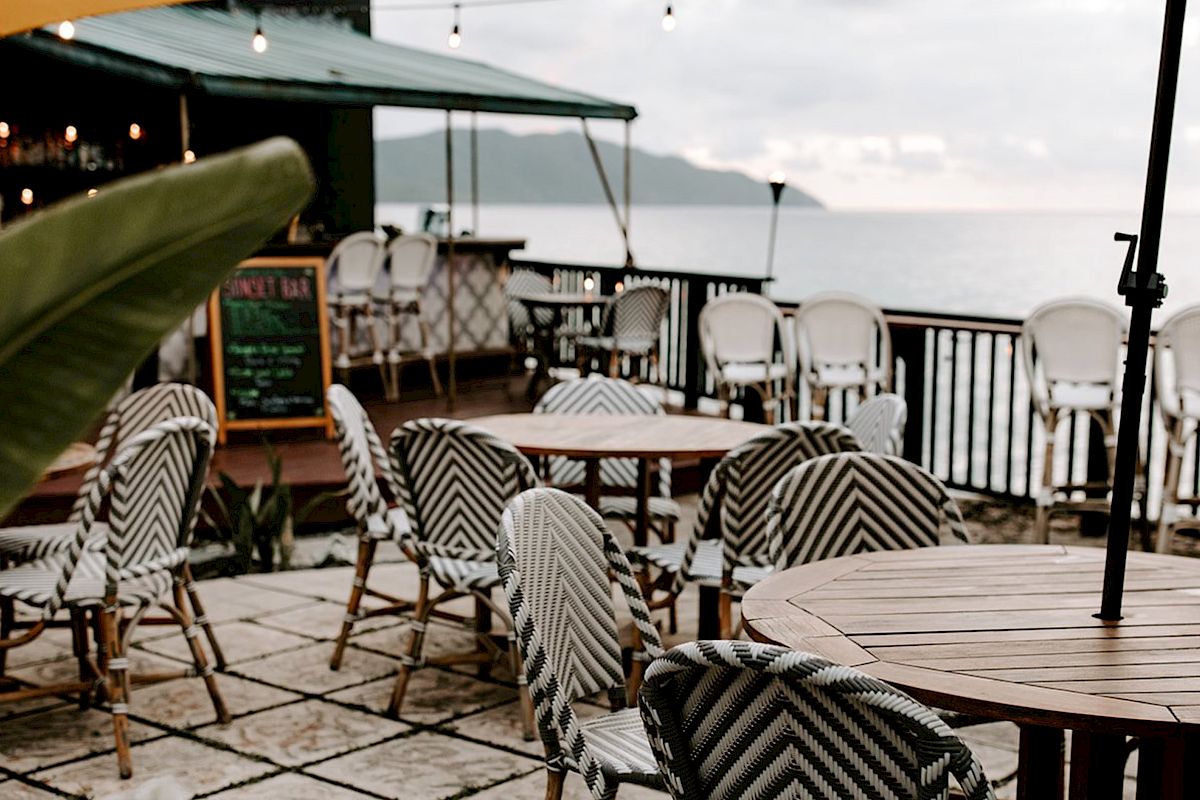 Outdoor seating with wooden tables and patterned chairs, overlooking the sea. There’s a bar area and string lights for ambiance.