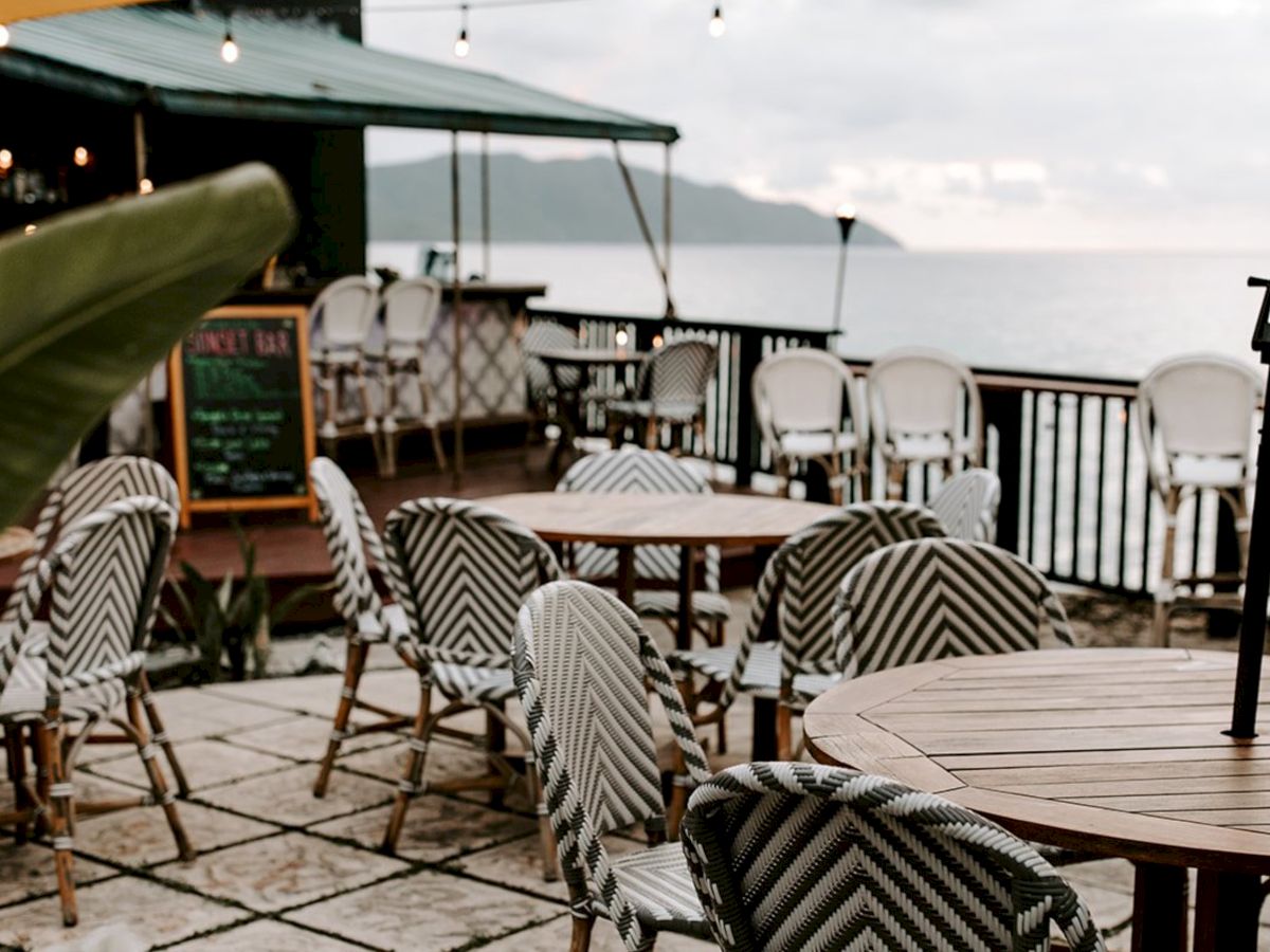 Outdoor seating with wooden tables and patterned chairs, overlooking the sea. There’s a bar area and string lights for ambiance.