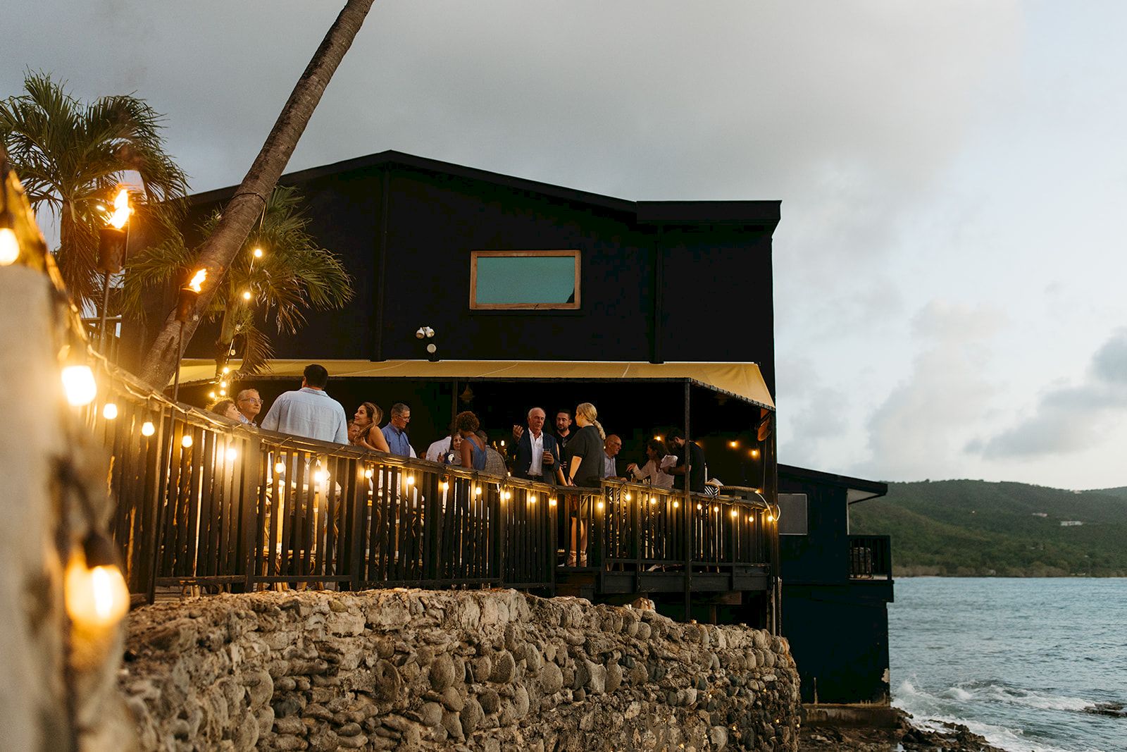 A group of people gather on a lit-up terrace by a coastal building during evening, with a scenic view of the sea and distant hills.
