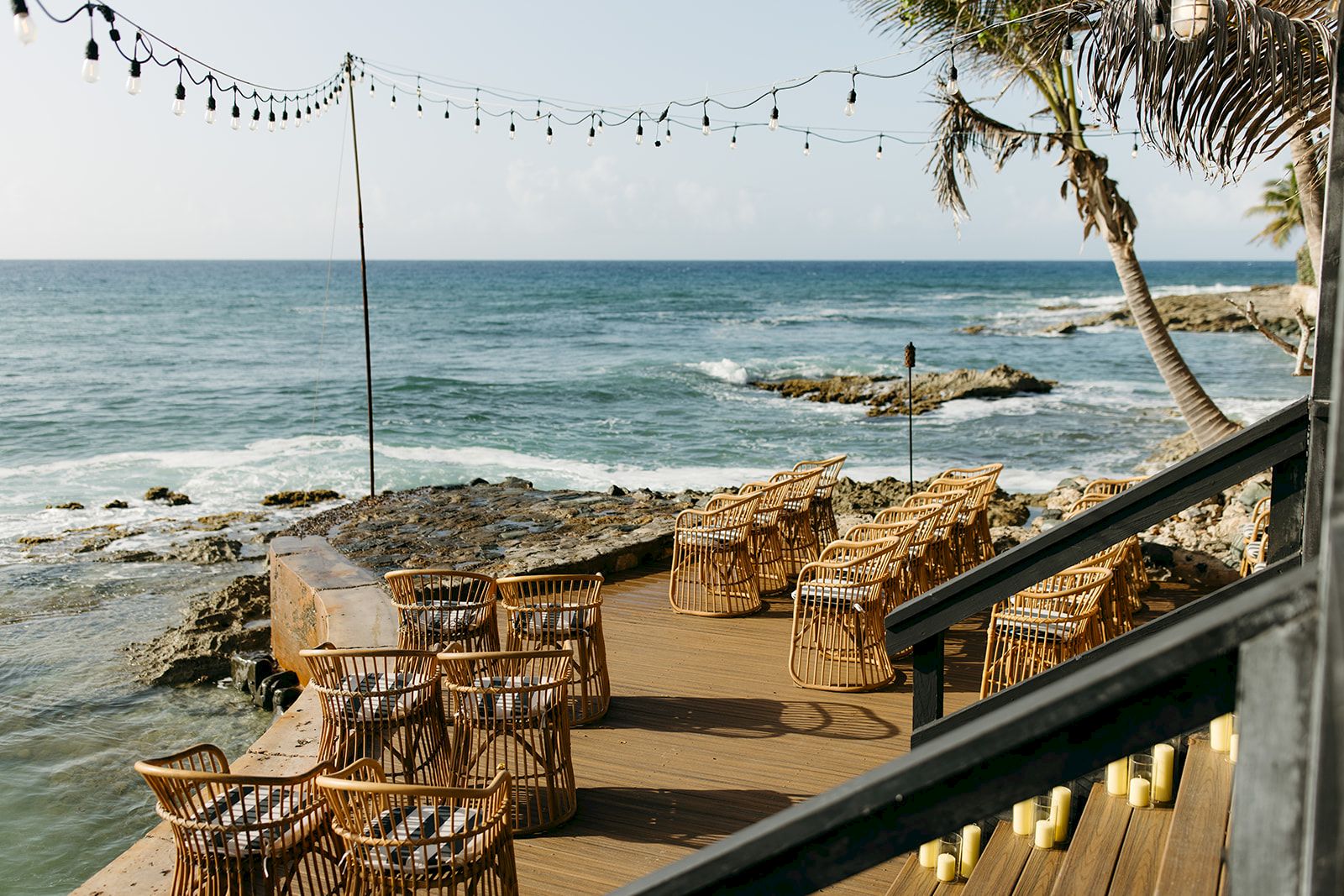 Oceanfront deck with wicker chairs and tables, surrounded by string lights and palm trees, overlooking a rocky shoreline under a clear sky.