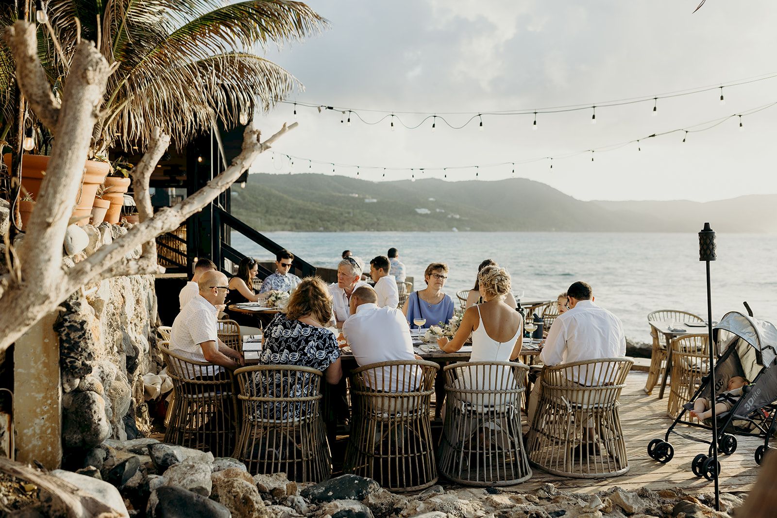 A group of people dining outdoors by the sea, seated at a long table with string lights overhead, surrounded by a scenic view.