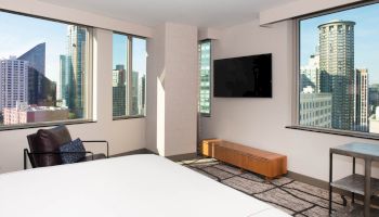 A modern hotel room with large windows, city view, television, chair, side table, and bed, offering a contemporary and inviting ambiance.