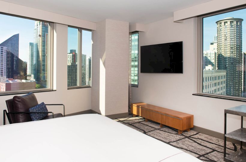 A modern hotel room with large windows, city view, television, chair, side table, and bed, offering a contemporary and inviting ambiance.