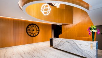 A modern lobby with marble flooring, wood paneling, a large clock, spherical lights, and a reception desk with flowers.