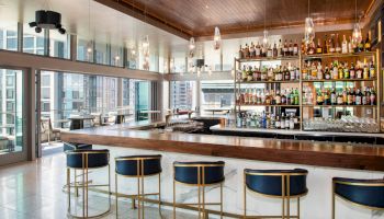 A stylish bar with high stools, a well-stocked shelf of bottles, and large windows showing a city view adorn the space.