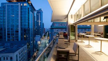 A modern cityscape at twilight shows tall buildings and a stylish balcony with tables and seating, glowing under ambient lighting.