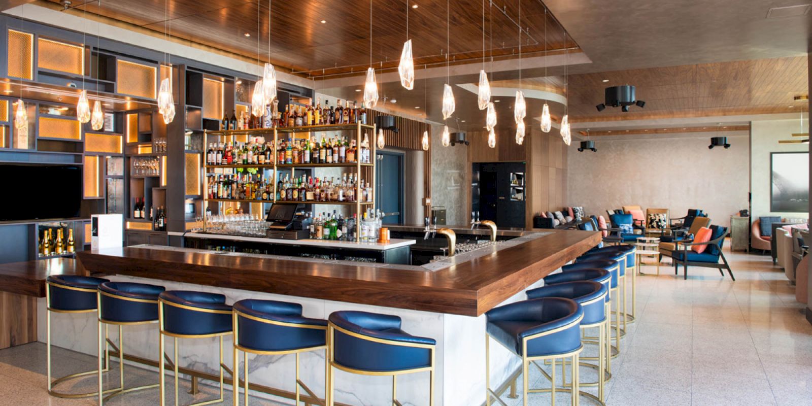 A modern bar with a wooden counter, blue barstools, pendant lights, and shelves stocked with bottles, in a chic lounge setting.