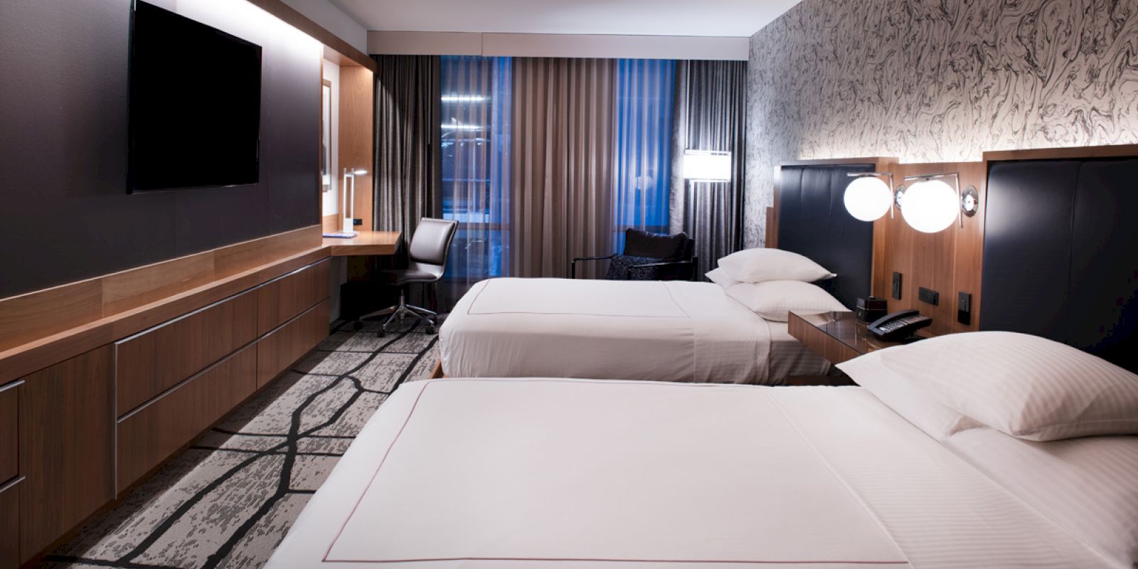 The image shows a modern hotel room with two beds, a desk, a TV, and patterned wallpaper, creating a cozy ambiance.