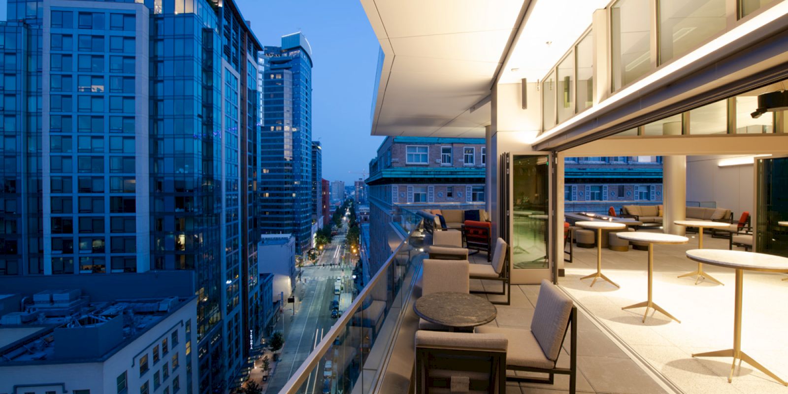 A modern urban balcony overlooks a city street, with seating and tables, adjacent to tall glass buildings under a twilight sky.