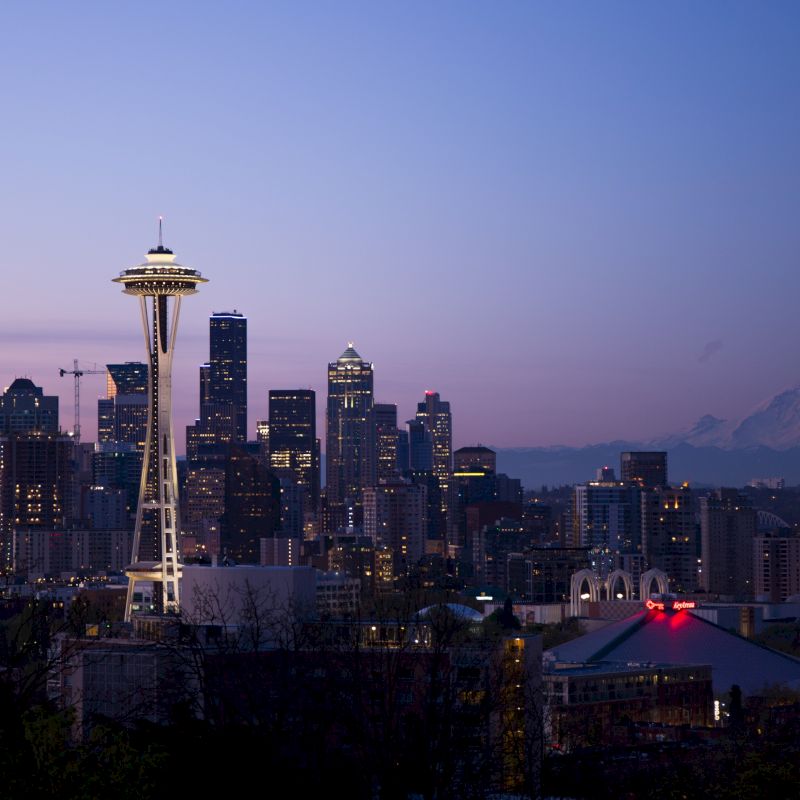 A city skyline at dusk with a distinctive tower and a mountain in the background, featuring illuminated buildings and a clear sky.