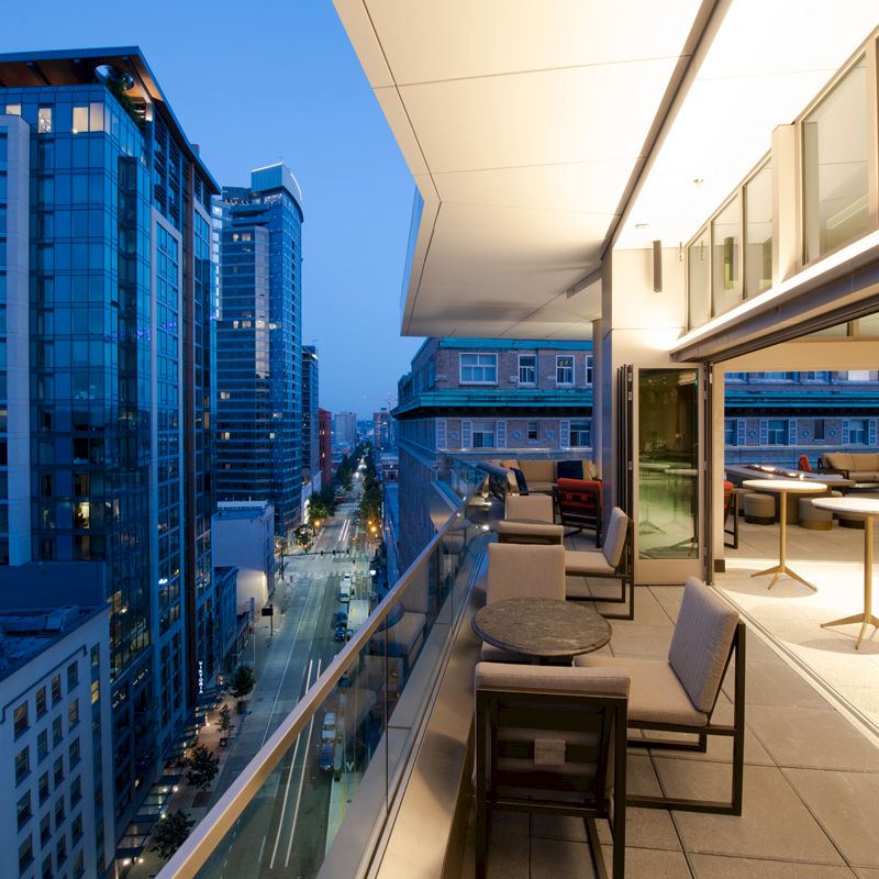 A modern urban scene with a cityscape view from a stylish rooftop terrace at dusk, featuring tables, chairs, and illuminated interiors.