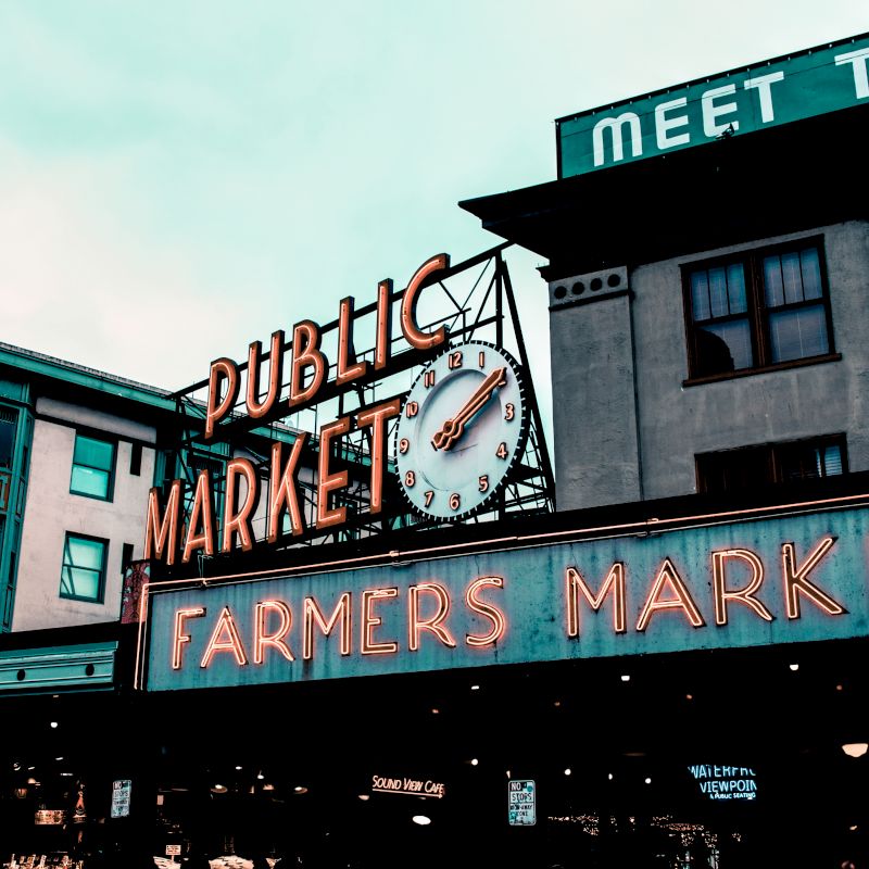 A historic public market sign with a clock, neon lights, and surrounding buildings against a cloudy sky are shown in this image.
