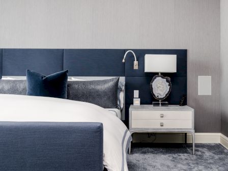 A modern bedroom features a navy and white color scheme, with a bed, nightstand, table lamp, and wall-mounted reading light.