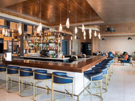 A modern bar with a square counter, high blue stools, overhead lights, and a fully stocked back wall in a contemporary lounge setting.