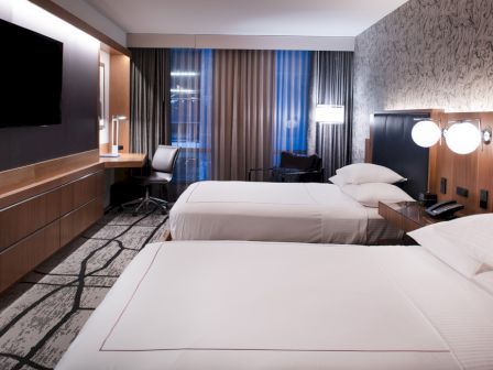A modern hotel room featuring two beds, wall-mounted TV, desk, and stylish decor with neutral tones, illuminated by soft lighting.