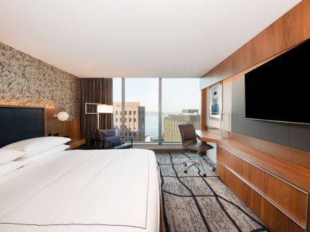 A modern hotel room with a large bed, desk, chair, and TV. There's a window with a cityscape and water view outside.