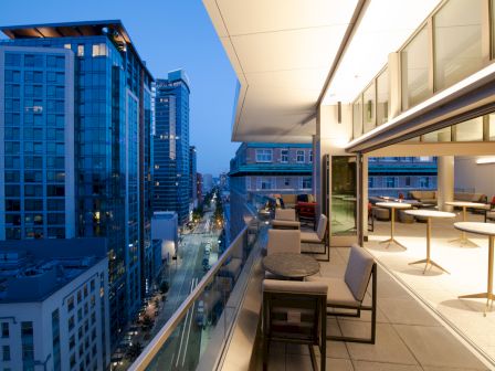 A modern cityscape at dusk, featuring high-rise buildings and a stylish balcony with tables and chairs overlooking the street below.