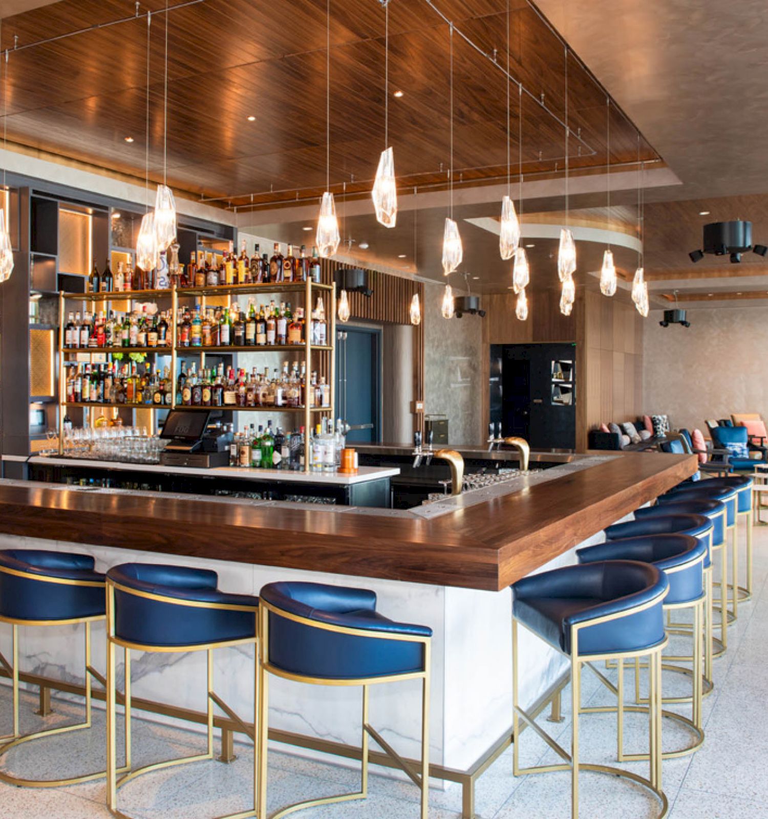 A modern bar with a wooden counter, blue chairs, hanging lights, and a well-stocked back bar, with seating and tables in the background.