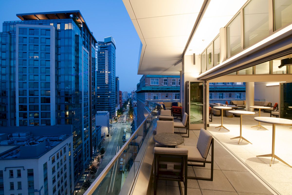 A modern cityscape view from a rooftop terrace featuring outdoor seating and tables, overlooking high-rise buildings at dusk.