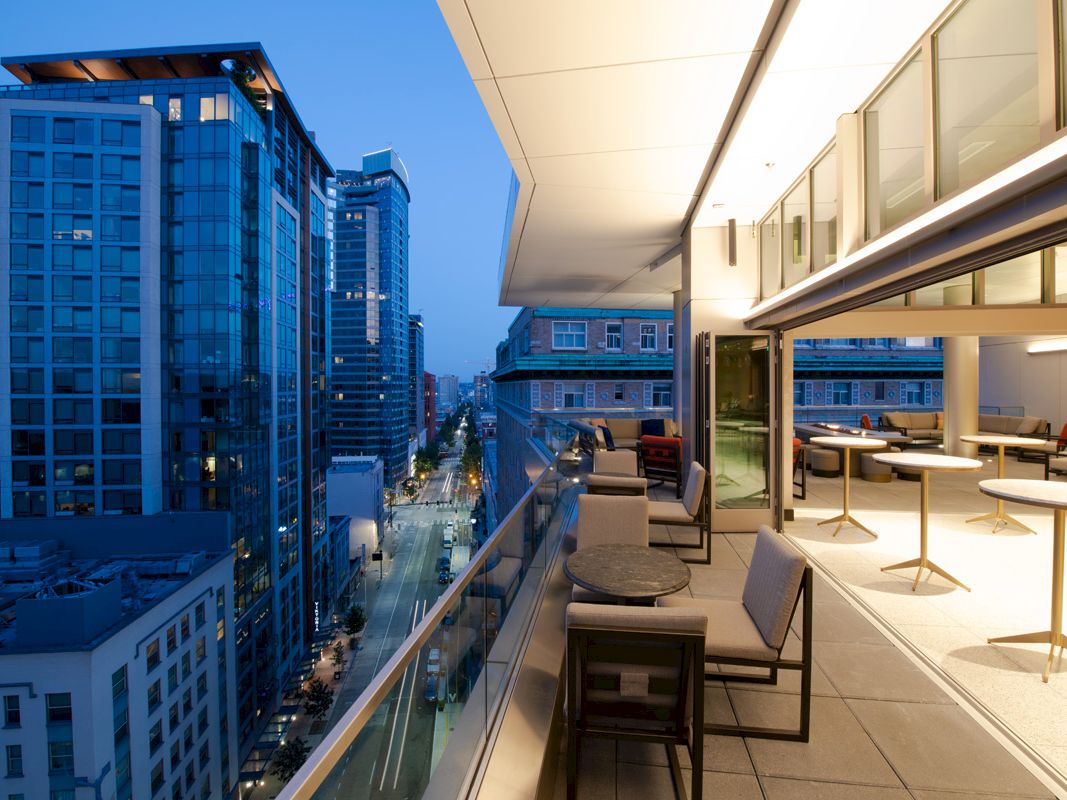 A modern cityscape view from a rooftop terrace featuring outdoor seating and tables, overlooking high-rise buildings at dusk.