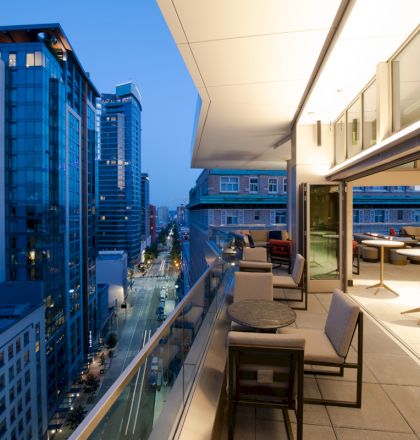 A modern rooftop terrace with seating overlooks a cityscape of tall buildings at dusk, featuring a blend of indoor and outdoor space.