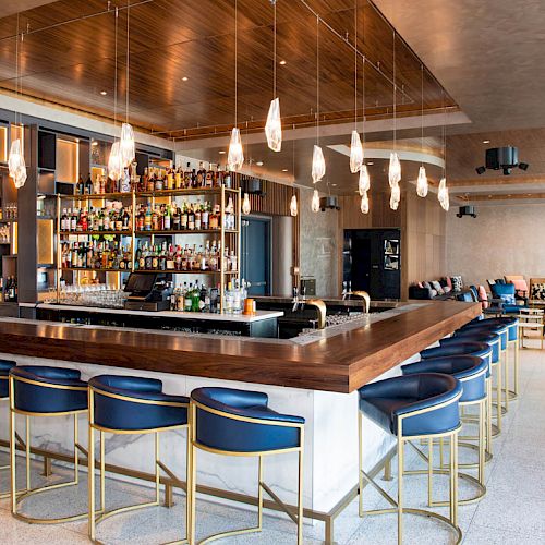 A modern bar with blue chairs, pendant lights, and a well-stocked bar area, featuring a lounge with comfortable seating in the background.