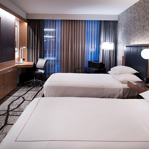 A modern hotel room with two beds, a flat-screen TV, a desk with a chair, and wallpaper accenting one wall.