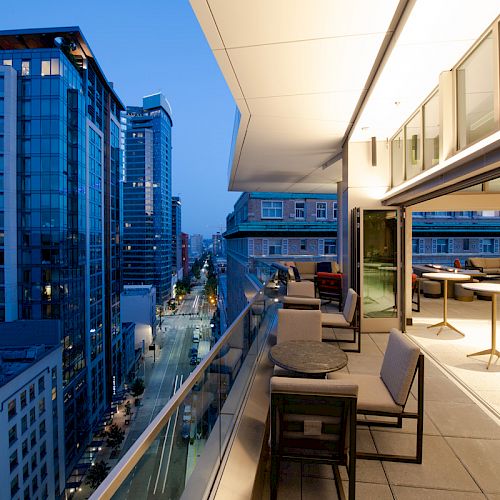 A modern balcony with seating overlooks a cityscape with tall buildings at dusk.