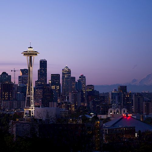 Seattle skyline at dusk with the Space Needle and Mount Rainier in the background, under a purple-blue sky.