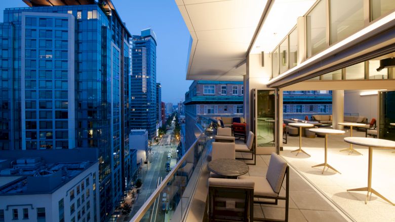The image shows a modern rooftop terrace with seating, city buildings, and a view of the street below during twilight.