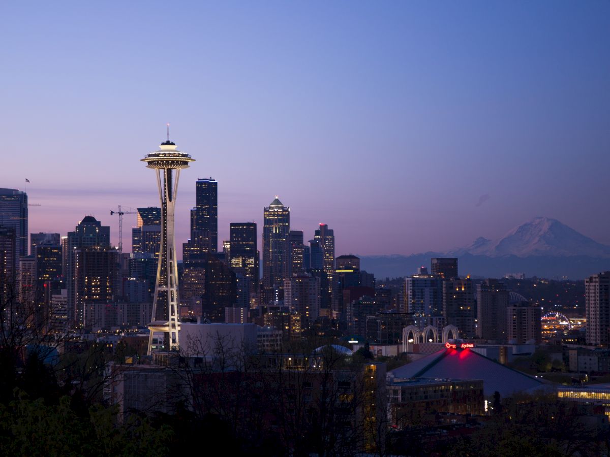 Seattle skyline at dusk with the Space Needle prominently featured, Mount Rainier in the background, and city lights illuminating.