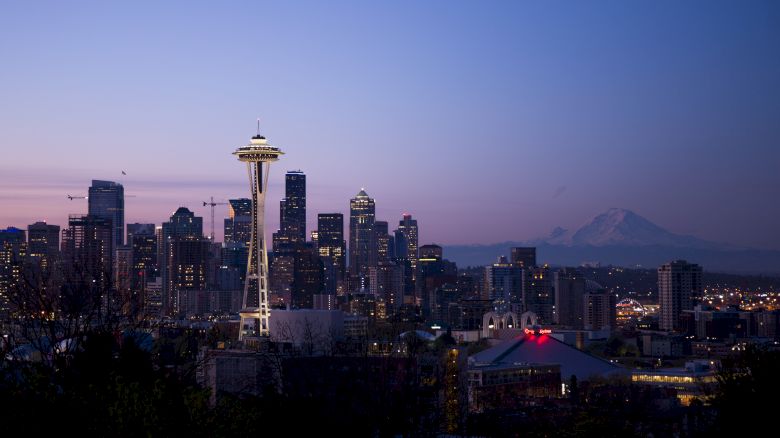 Seattle skyline at dusk with the Space Needle prominently featured, Mount Rainier in the background, and city lights illuminating.