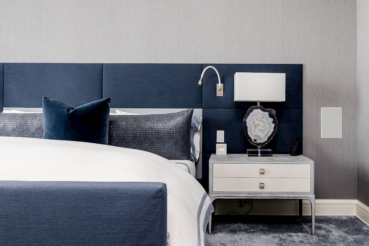 A modern bedroom with a navy headboard, white bedding, nightstand with a lamp, and decorative items.