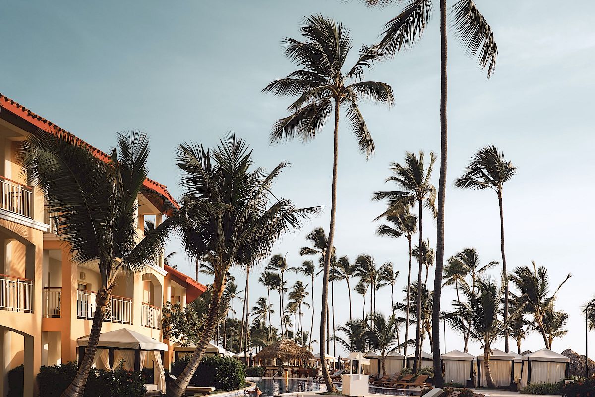 A resort poolside area with lounge chairs, palm trees, and a building in the background under a clear sky.