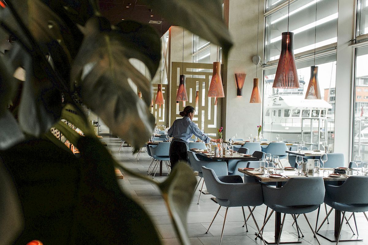 A restaurant interior with a person setting tables, surrounded by modern chairs and pendant lights, viewed through leafy plants.