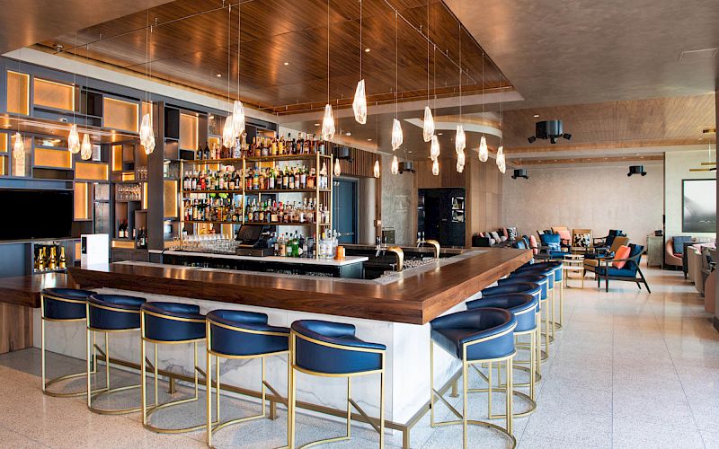 A stylish bar with a central countertop, surrounded by blue stools, modern lighting, and shelves filled with various bottles.