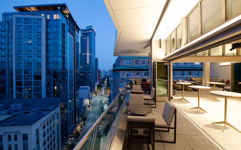 A modern cityscape at twilight with high-rise buildings and a terrace featuring tables and chairs, overlooking a street.